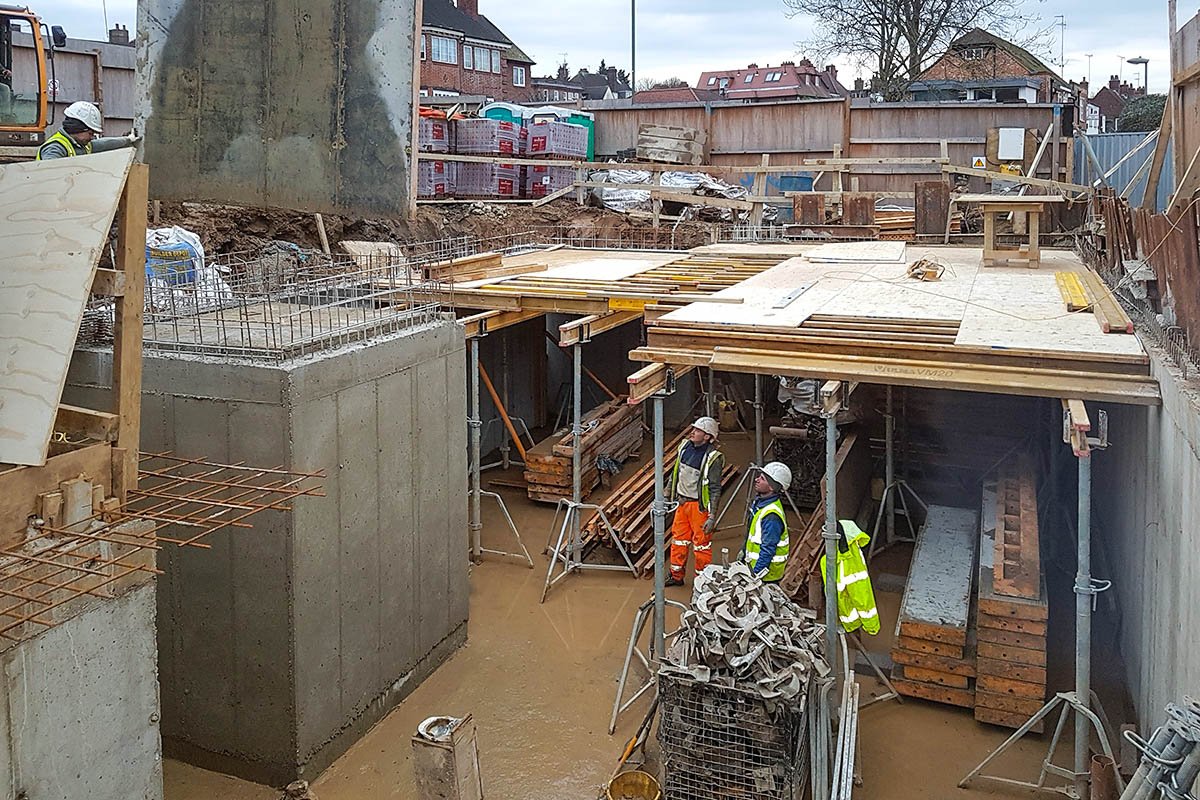 Hodford Road workmen in high-viz working within new concrete basement lifting temporary formwork into place with beams and acro props